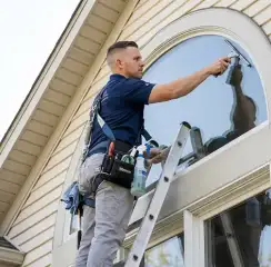 Professional window cleaner on a ladder using a squeegee to clean an exterior arched window, Rochester, NY.